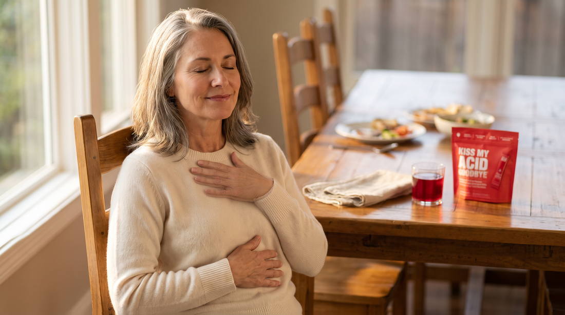 A woman in her fifties sits at a dining table with her eyes closed, one hand on her chest and one on her belly, taking a slow breath; a glass of red KMAG drink and a KMAG pouch sit on the table behind her