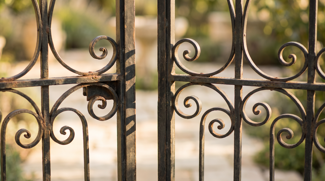Close-up editorial photograph of a weathered wrought-iron garden gate left slightly ajar in warm morning light